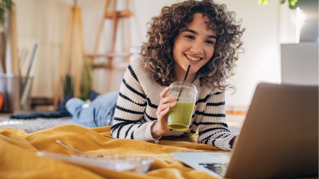woman drinking a green smoothie while looking at her laptop