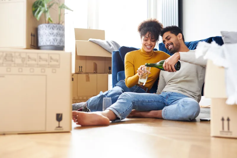 man and woman sitting on the floor, pouring a beverage while surrounded by boxes.