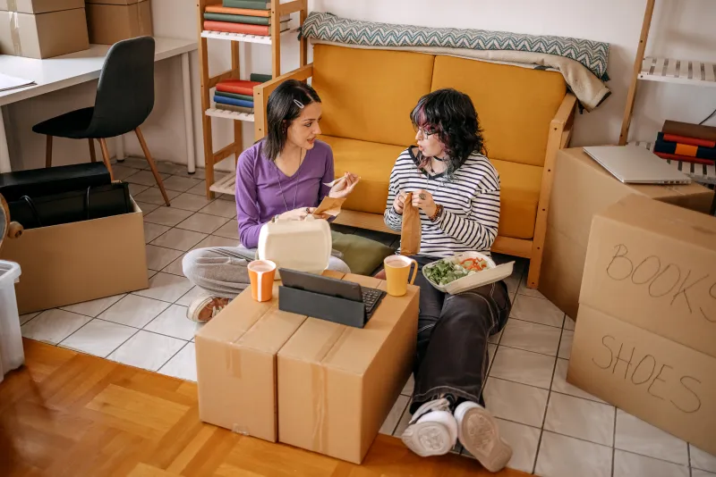 two roommates sitting among boxes eating take-out food