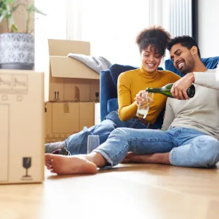 man and woman sitting on the floor, pouring a beverage while surrounded by boxes.