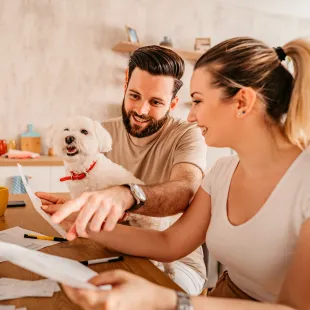 A couple sitting at a computer with paperwork. The man has a small white dog on his lap.