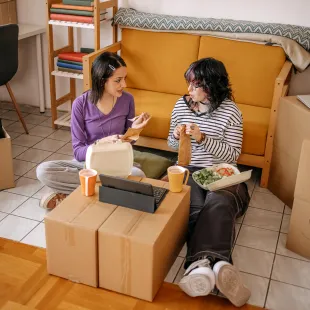 two roommates sitting among boxes eating take-out food